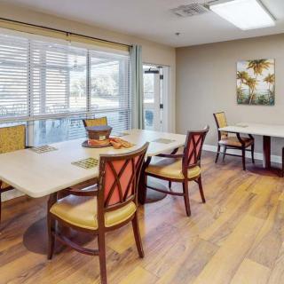 A well-lit dining room with wooden floors, featuring several tables and chairs, and bright natural light from large windows.