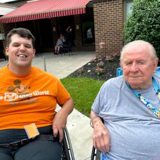 Two individuals enjoying a sunny day outdoors, both smiling and seated in wheelchairs.