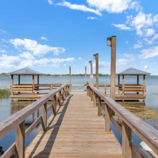 A scenic view of a wooden pier extending into a serene lake, flanked by gazebos under a bright blue sky with scattered clouds.