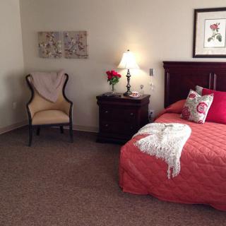Cozy bedroom with a bed draped in a red quilt, an elegant chair, and a warmly lit nightstand.