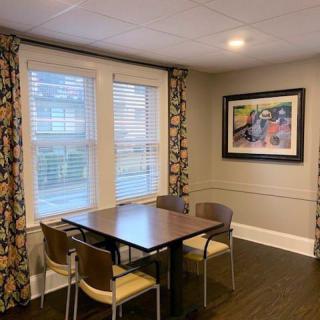 A modern dining area with a wooden table, four chairs, large windows with patterned curtains, and framed artwork on the wall.