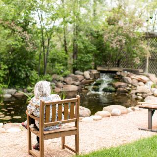 A person sits on a bench by a pond with a small waterfall in a tranquil park setting.
