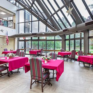 A bright and airy dining area featuring tables with red tablecloths, surrounded by large windows that let in natural light.