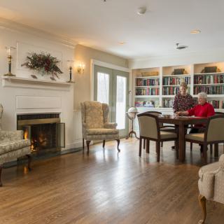 A cozy living room with a fireplace, bookshelves, and two people sitting at a table.