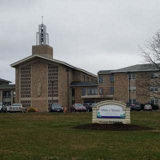 A modern church building with a prominent tower and an adjacent parking lot under an overcast sky.