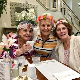 Three friends enjoying a celebration, wearing floral crowns and smiling together at a dining table.