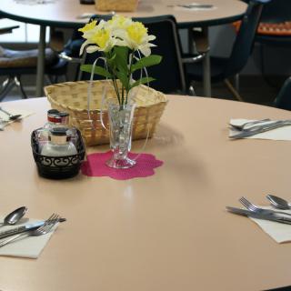 A dining table is set with silverware, a bottle of syrup, and a vase of flowers.