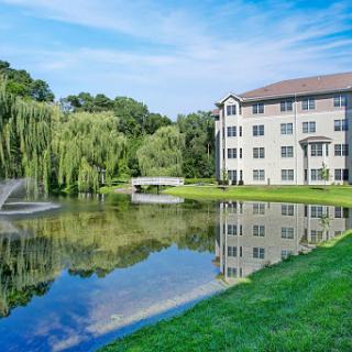 A serene view of a building by a pond, featuring lush greenery and a fountain.
