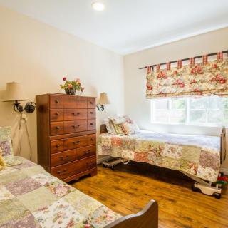A cozy bedroom with twin beds featuring quilted bedspreads, a wooden dresser, and floral curtains over the window.