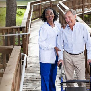 A nurse assisting an elderly man using a walker on a wooden path outdoors.