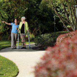 A caregiver assists an elderly woman using a walker as they stroll along a garden path.