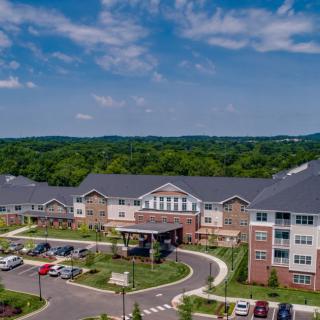 An aerial view of a large building complex with a parking lot, surrounded by trees under a partly cloudy sky.