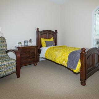 A cozy bedroom with a wooden single bed featuring yellow and gray bedding, adjacent to a nightstand and an upholstered chair.