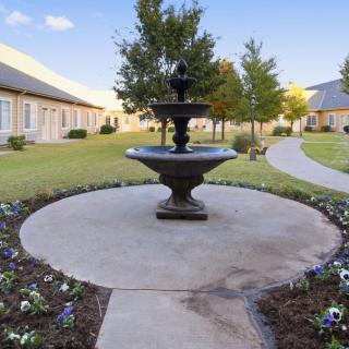 A serene courtyard with a central fountain, surrounded by landscaped gardens and winding pathways, flanked by buildings.