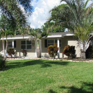 A single-story house surrounded by palm trees and a well-maintained lawn under a clear blue sky.
