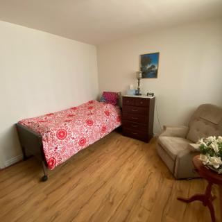 A cozy bedroom features a single bed with a floral bedspread, an armchair, and a dresser topped with a lamp and decorative items.