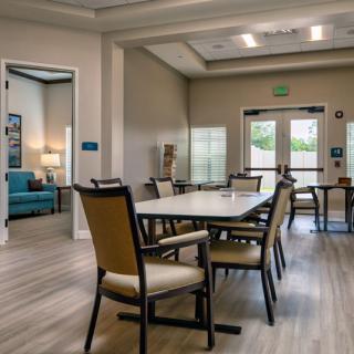A well-lit community dining area with tables and chairs, featuring wooden flooring.