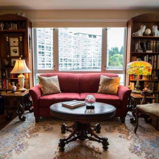 A cozy living room with a red sofa, surrounded by bookshelves and illuminated by soft natural light through the window.