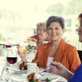 An elderly woman enjoying a meal and wine at an outdoor social gathering.