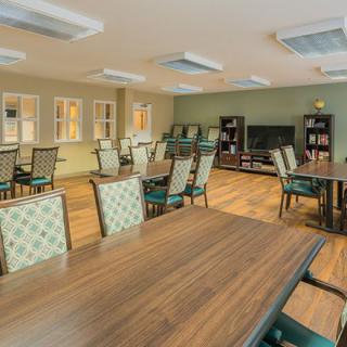 A spacious dining room with multiple tables and chairs, featuring a wooden floor and a television opposite bookshelves.
