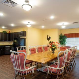 A cozy dining area featuring a long wooden table surrounded by cushioned chairs, adjacent to a kitchen space with dark cabinetry.