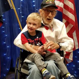 A heartwarming moment shared between an elderly man in a wheelchair and a young boy, both smiling against a backdrop of American flags.