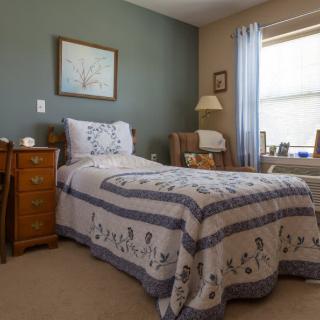 A cozy bedroom with a single bed and quilt, wooden furniture, and a window with light blue curtains.