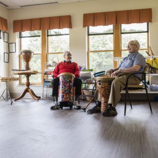 Two elderly individuals playing drums indoors near large windows.