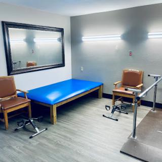 A view of a physical therapy treatment room featuring a blue table, two chairs, and various rehabilitation equipment.