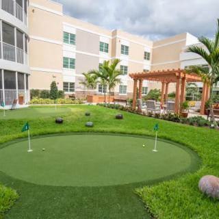 A neatly landscaped outdoor area with a small golf putting green and a pergola, adjacent to a modern building.