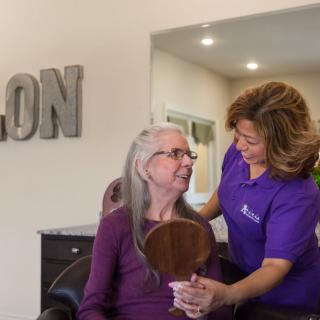 A stylist assists an elderly woman in a salon, holding a mirror to show her hairstyle.