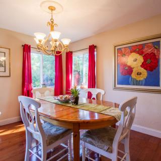 A cozy dining room featuring a wooden table, chairs, chandelier lighting, vibrant red curtains, and colorful artwork.