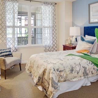 A cozy bedroom with a neatly made bed, soft lighting, and a large window letting in natural light.