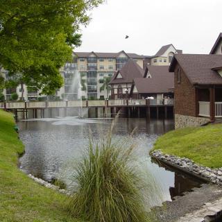 A scenic view of buildings by a tranquil lake with a fountain and surrounding greenery.