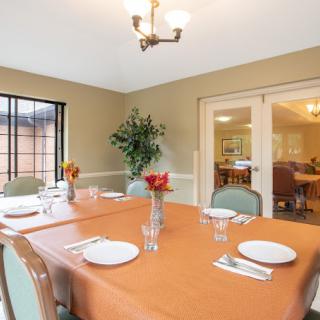 A neatly arranged dining room with an orange tablecloth, set with plates and cutlery, featuring a vase of flowers in the center.