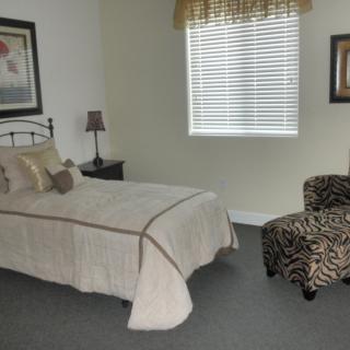 A cozy bedroom with a neatly made bed, a patterned chair, and soft natural light coming through the window.