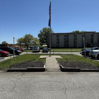 A clear day showcasing a flag in front of a parking area, flanked by neatly trimmed grass and a distant building.