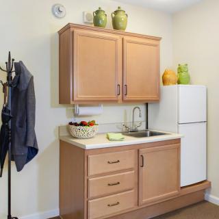 A small kitchen with wooden cabinets, a sink, and a white refrigerator. Decorative green vases are displayed above the cabinetry alongside a fruit bowl on the counter.