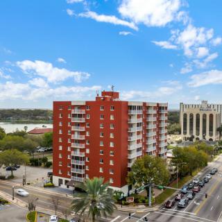 A red brick apartment building situated at a busy intersection in an urban area, with surrounding greenery and a clear blue sky.