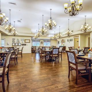 An elegant dining room with wooden floors, furnished with multiple tables and chairs, illuminated by ornate chandeliers.