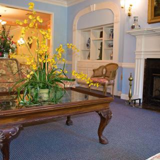 A cozy living room with a traditional style, featuring a glass coffee table adorned with yellow flowers and a classic fireplace.