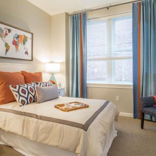 A modern and cozy bedroom with stylish decor, featuring a neatly made bed with colorful pillows, large windows with elegant curtains, and a comfortable armchair.