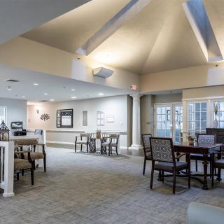 A spacious and well-lit dining area with multiple tables and chairs, featuring a carpeted floor and large windows.