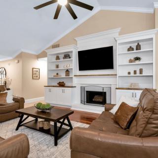 Cozy living room featuring leather sofas, a central coffee table, built-in shelves around a fireplace and TV, with a ceiling fan overhead.