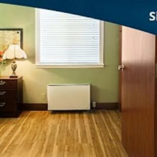A cozy bedroom with wooden flooring, featuring a nightstand with a lamp, a window with blinds, and an open door.