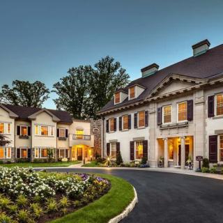 A grand luxury mansion illuminated during the evening with landscaped gardens and a circular driveway.