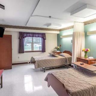 A view of a hospital patient room featuring two beds, a window with curtains, and basic furniture.