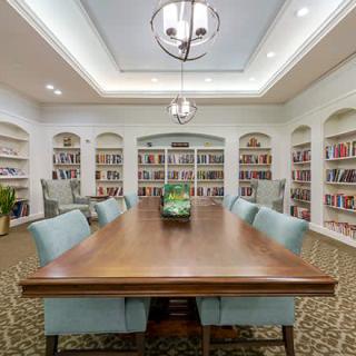 A well-lit library with a large wooden table surrounded by chairs, featuring bookshelves filled with books.