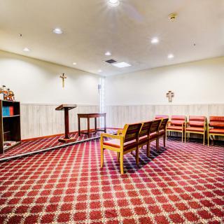 A small chapel interior with chairs arranged on a patterned carpet, facing a pulpit and religious symbols on the walls.