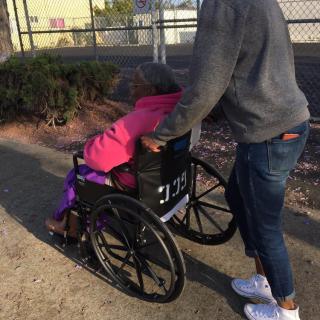 A person assists an elderly individual in a wheelchair outdoors.
